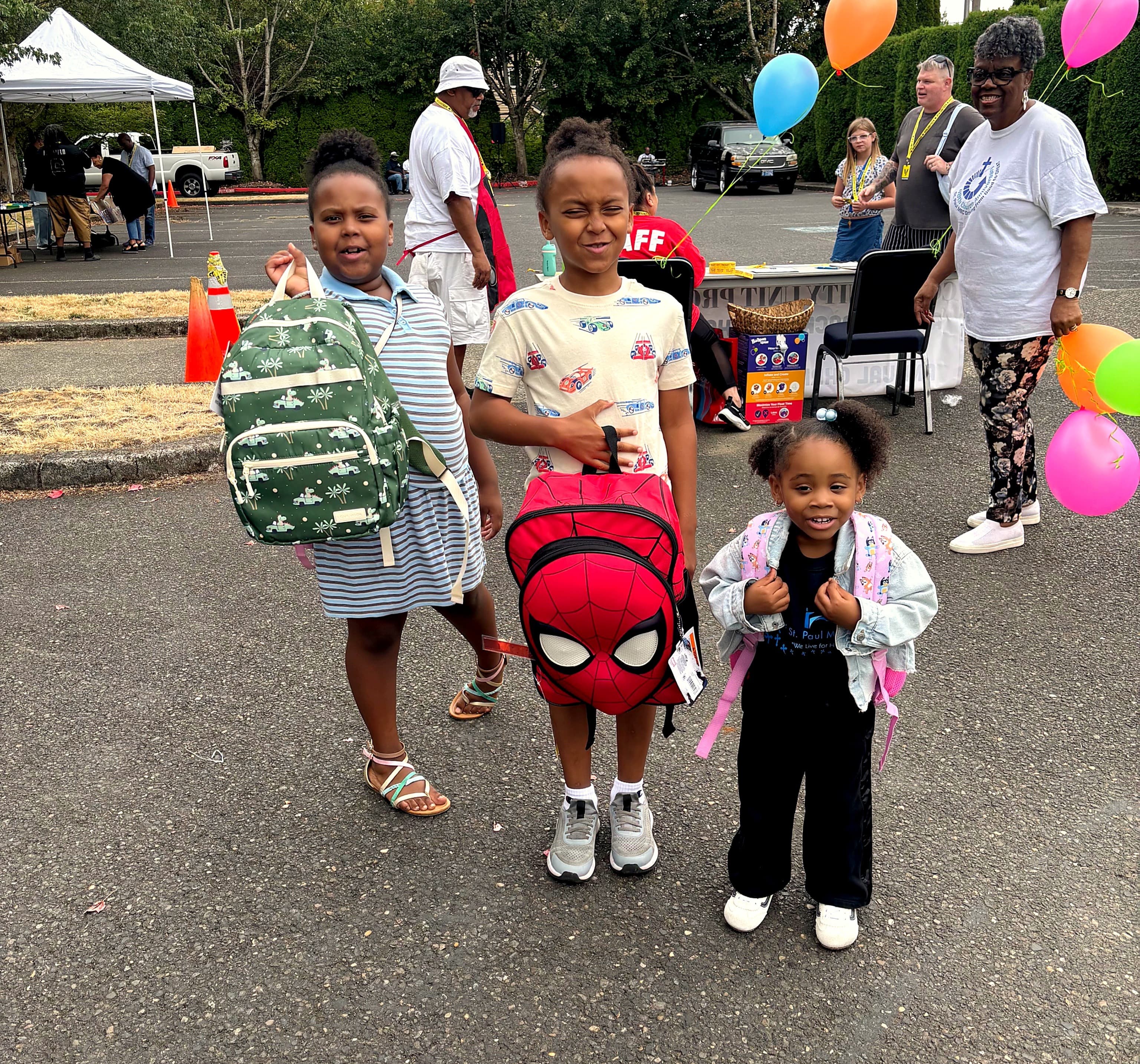 Three kids smiling and holding new backpacks at a giveaway event