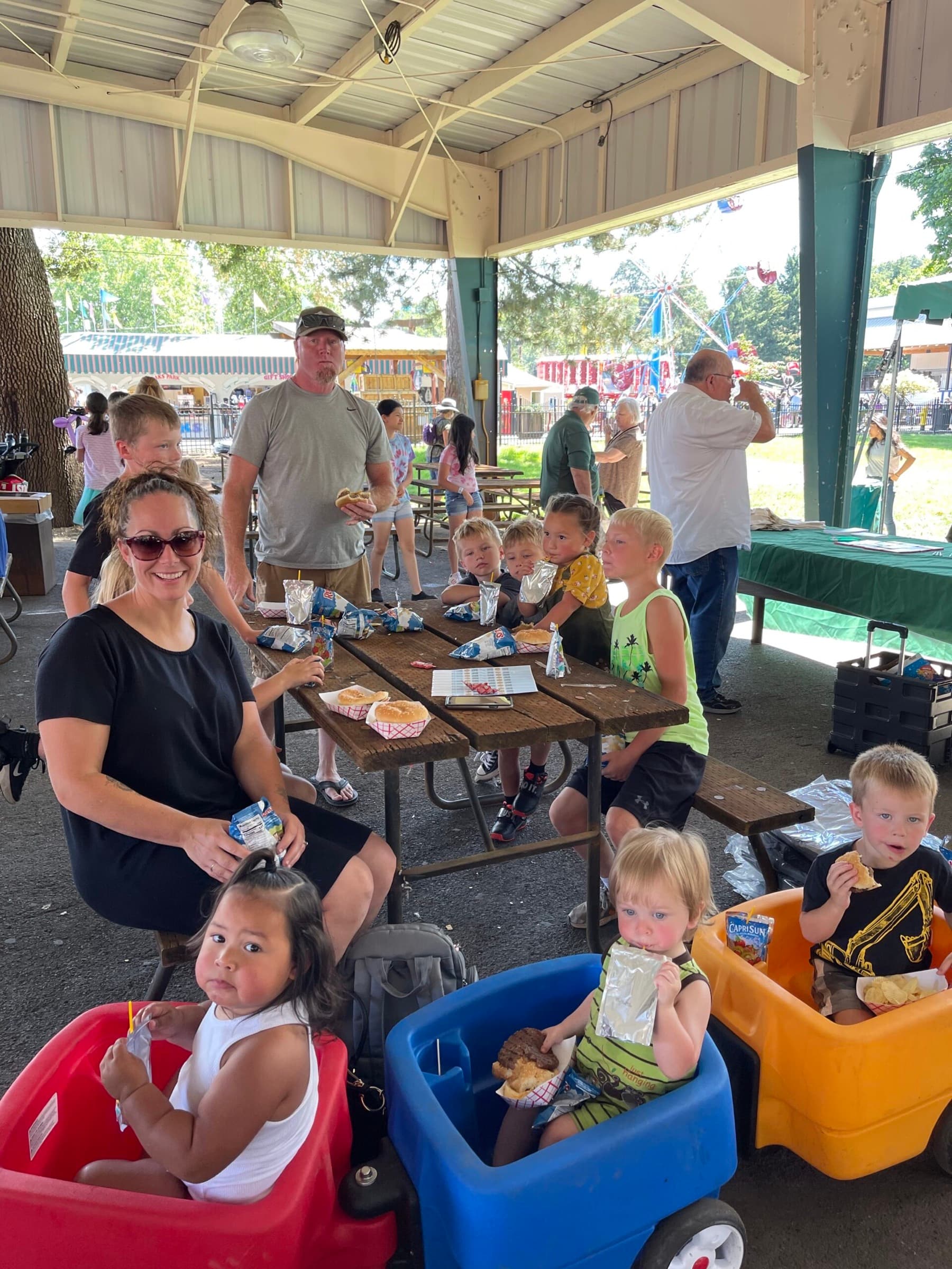 Families and kids eating hot dogs at a picnic table under the pavilion at Oaks Park Day, with carnival rides in the background